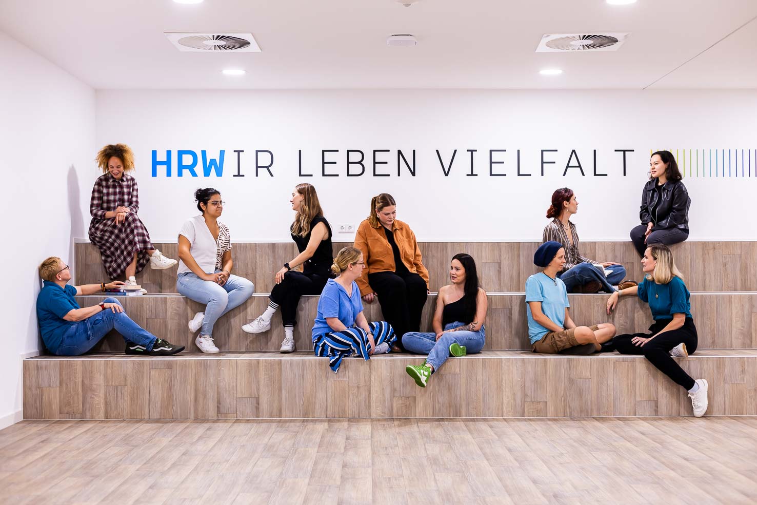 11 weiblich gelesene Personen sitzen auf Stufen vor einer Wand auf der "HRWIR LEBEN VIELFALT" steht